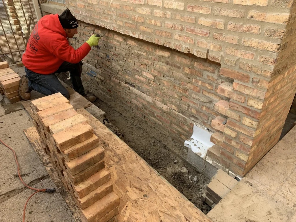 Mason in red jacket repairs damaged brick wall with new bricks.
