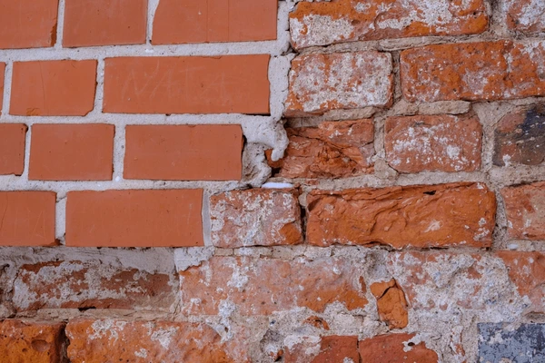 Close-up view of a weathered brick wall with intact orange bricks on the left and damaged, crumbling red bricks with white mortar on the right.