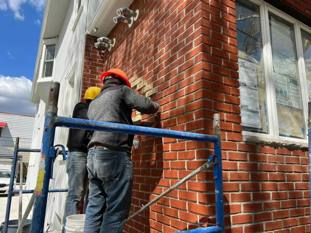 Two workers in hard hats stand on scaffolding, installing brickwork on the side of a building next to a window.