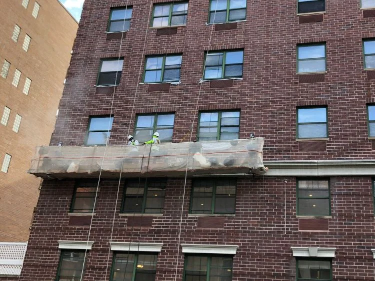 Two workers in hard hats are on a suspended scaffolding platform cleaning the exterior of a brick building.