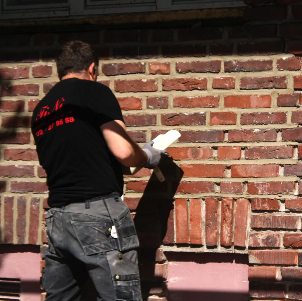 A bricklayer in a black shirt and gray work pants uses a trowel to apply mortar to a brick wall.