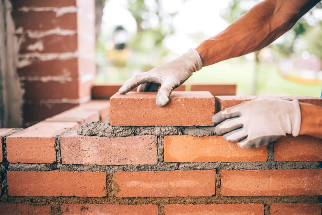 A gloved hand carefully places a brick atop a row of freshly laid bricks, with mortar visible between them. Soft focus on greenery behind.