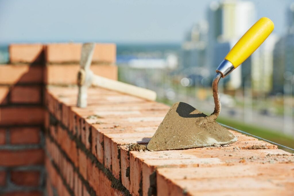 Trowel and hammer on a brick wall under construction with industrial buildings in the background.