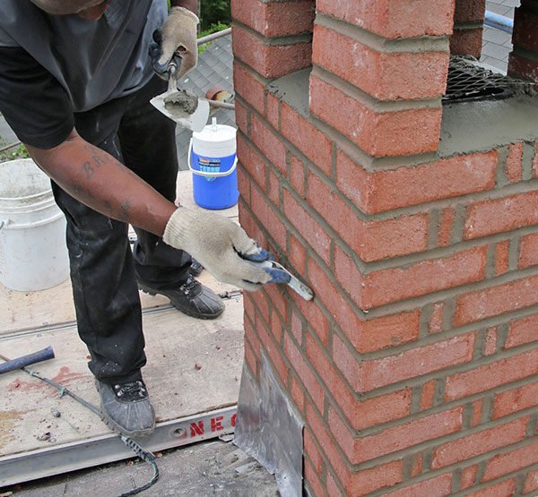 A worker in gloves applies mortar to a brick chimney using a trowel, standing on a platform with construction tools nearby.