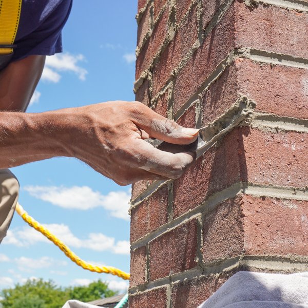 A mason uses a trowel to apply mortar to a brick chimney under a blue sky.