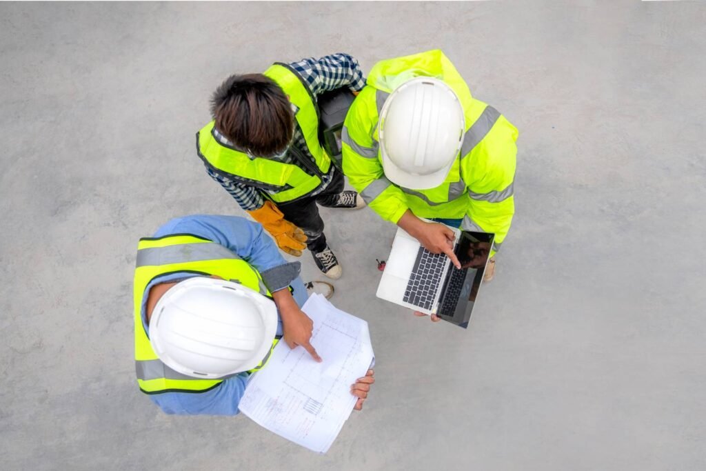 Construction workers in safety vests and hard hats reviewing blueprints and laptop on site planning project details.