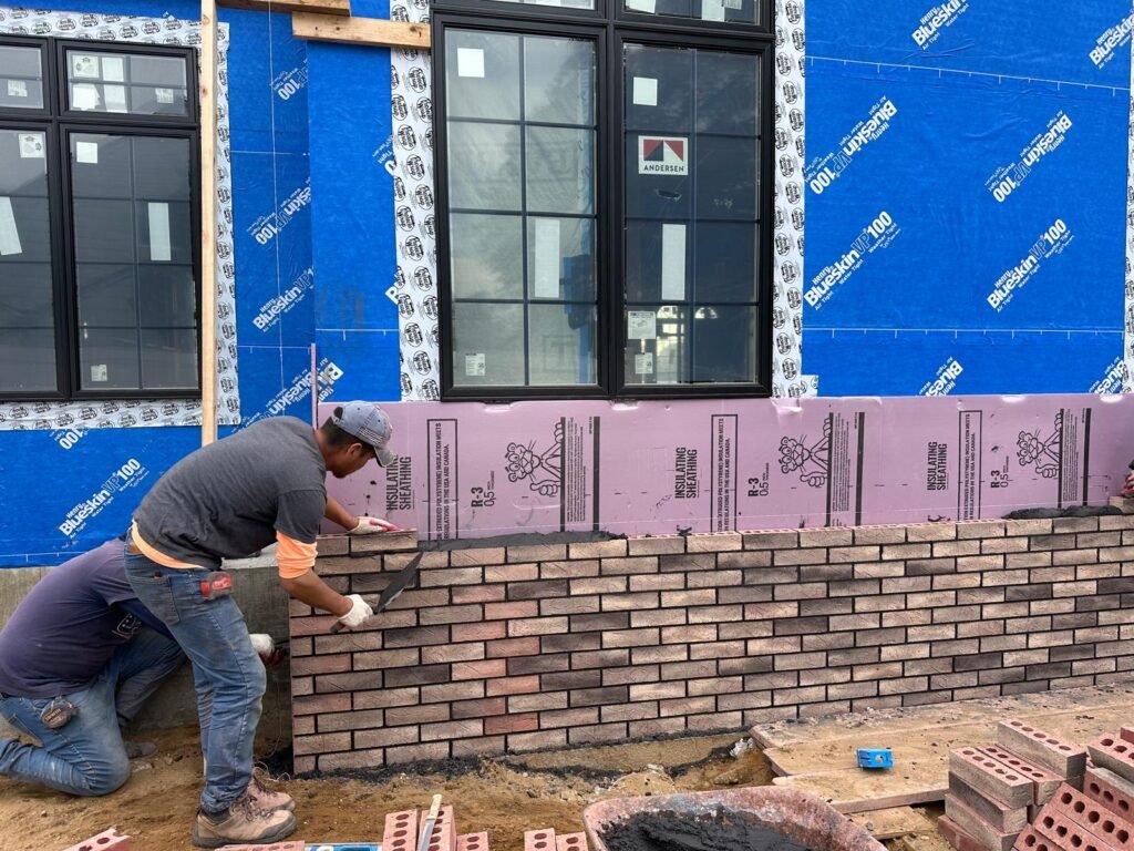 Two construction workers lay bricks on a building's exterior, with blue insulation and large windows visible in the background.