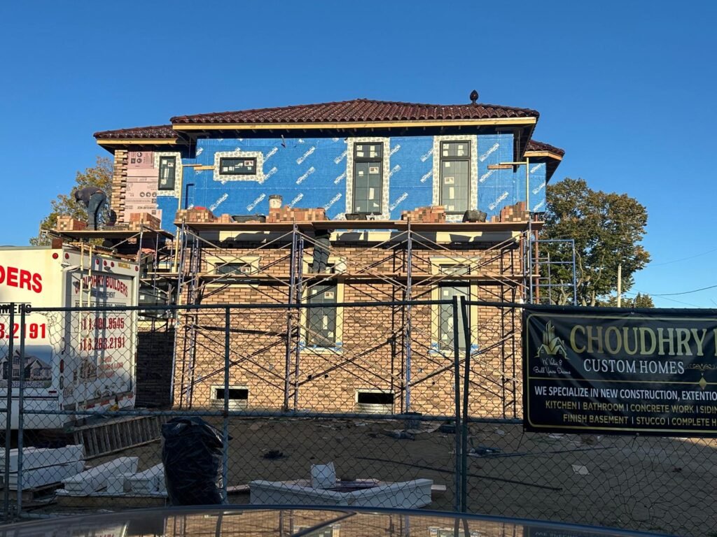 Construction workers install brick siding on a house wrapped in blue house wrap, with scaffolding and a company banner visible.