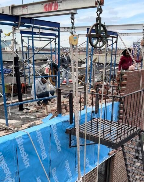 Construction workers are seen on a rooftop with scaffolding, pulleys, and blue building wrap.