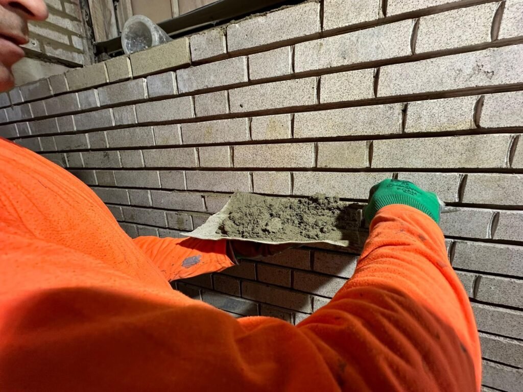 A construction worker in an orange shirt holds a trowel with mortar, preparing to apply it to a brick wall.