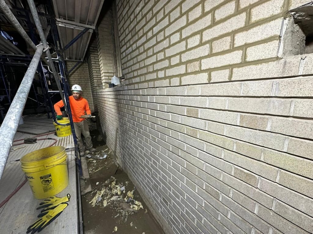 A construction worker in an orange shirt and white hard hat stands near a freshly mortared brick wall, with scaffolding and yellow buckets on the left.