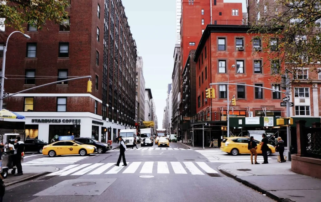 Pedestrians cross a street lined with brick buildings and yellow taxis in New York City.