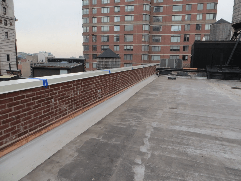 Rooftop view with brick parapet wall, flat roof, and distant cityscape including a tall brick building and water towers.
