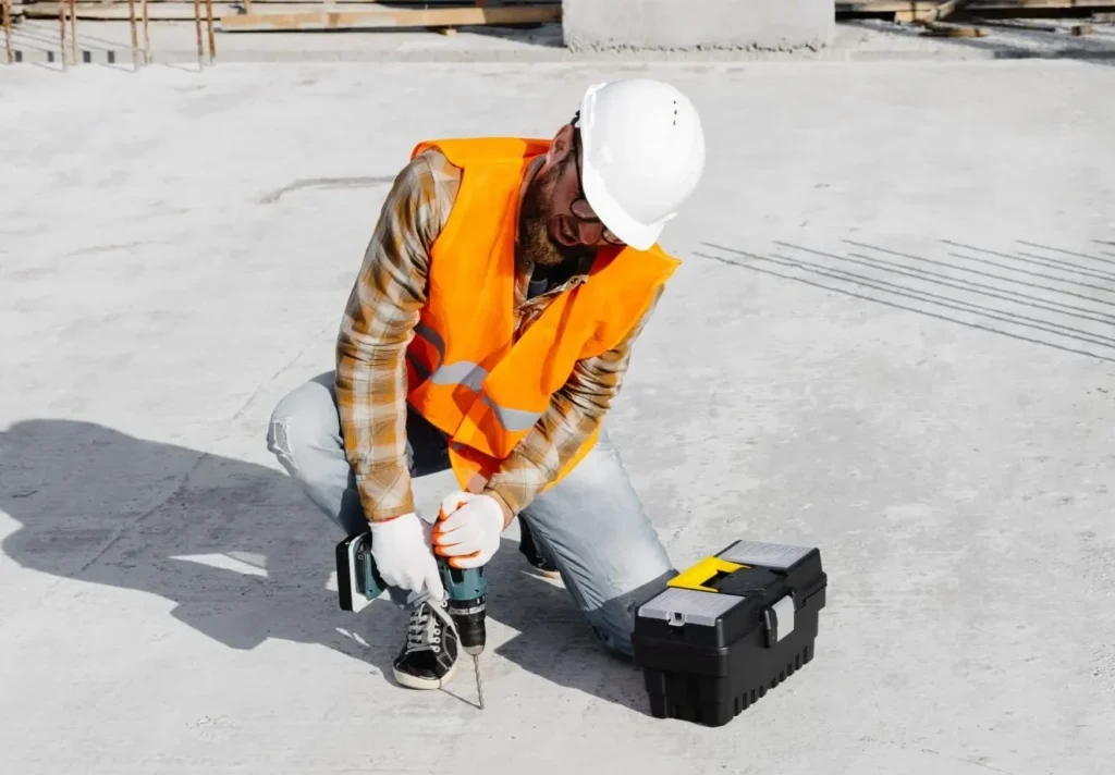 A construction worker in a neon vest kneels on a concrete surface, using a power drill beside a toolbox.