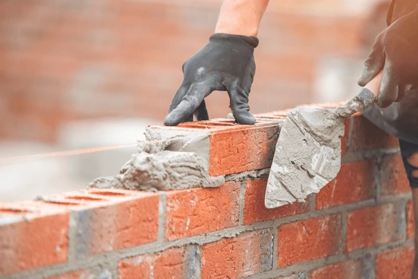 Bricklayer wearing gloves places a brick onto a mortar-covered wall with a trowel.
