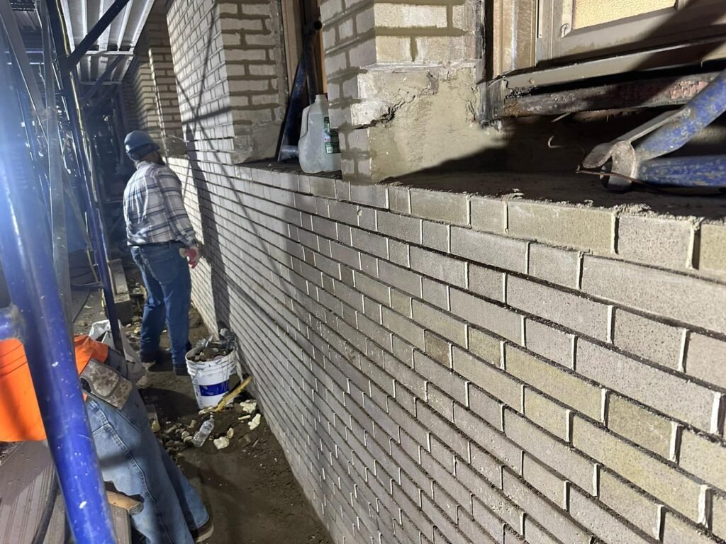 A construction worker in an blue shirt and white hard hat stands near a freshly mortared brick wall, with scaffolding and yellow buckets on the left.